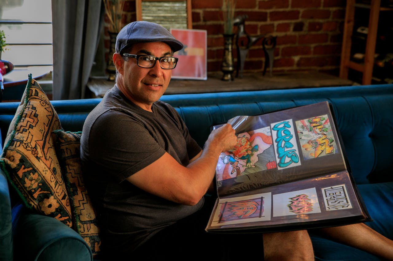 The Art of Drawing Readers In: Your attractive post title goes here Man in hat and glasses proudly displaying his colorful graffiti art portfolio on a couch.